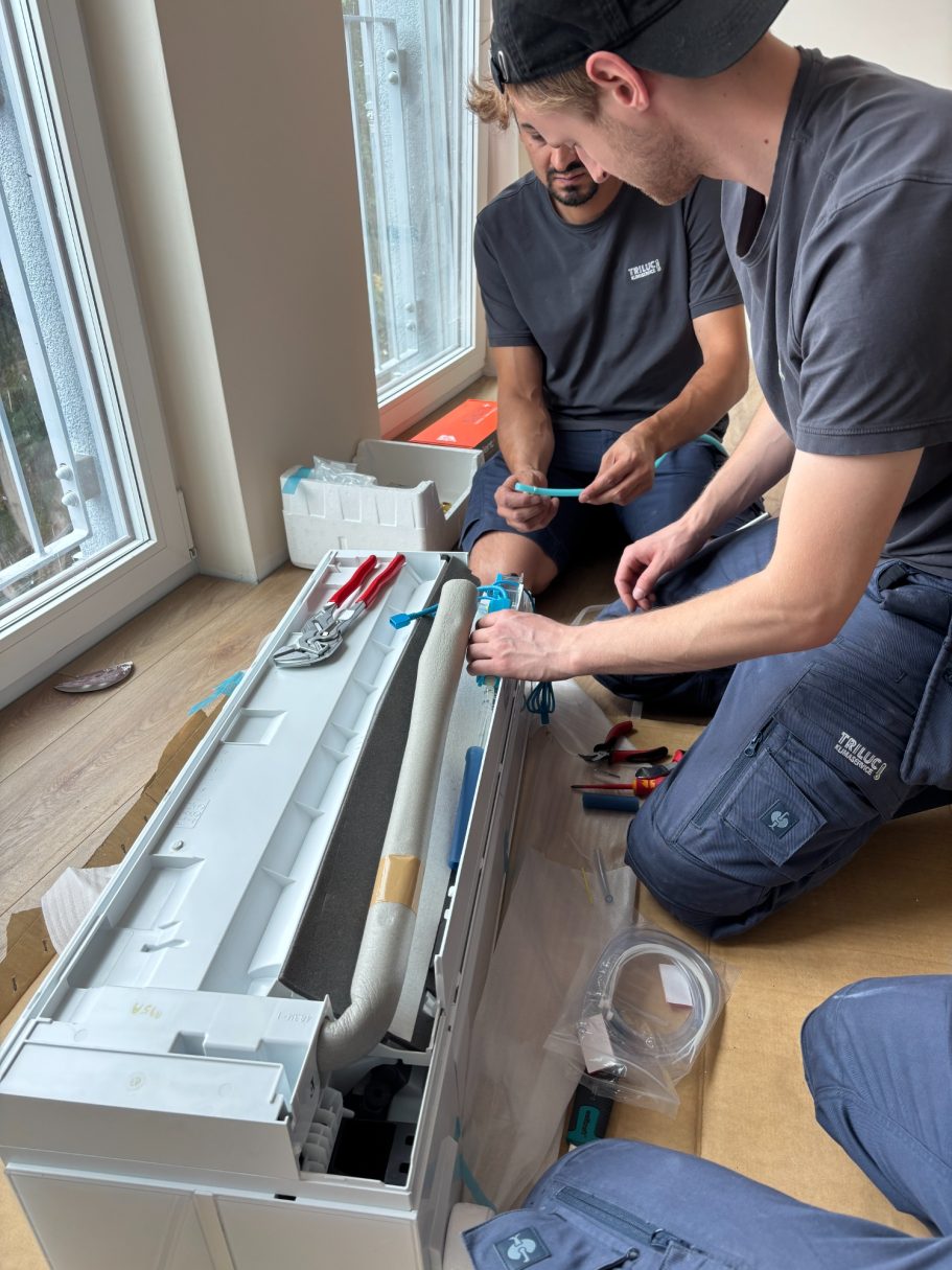 Drei TRILUC Kältetechniker arbeiten gemeinsam und konzentriert an einer zu installierenden Klimaanlage, Werkzeuge liegen auf dem Boden.