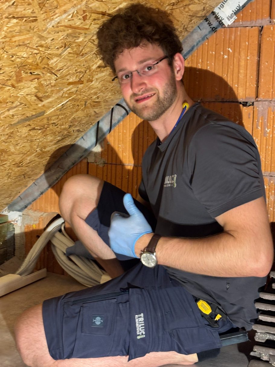 Junger TRILUC Kältetechniker, der in einem Dachboden anerkennend Daumen hoch zeigt.