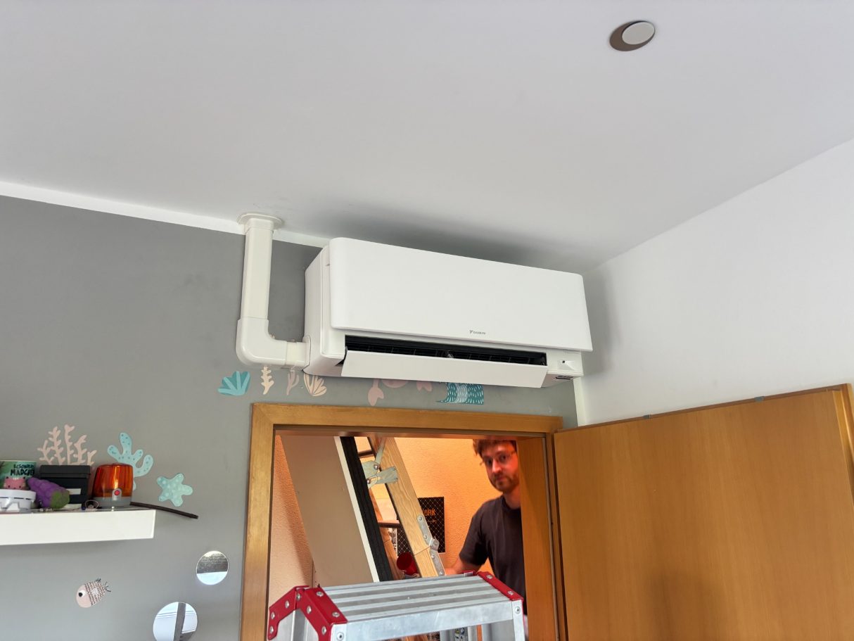 Weißes Daikin Klimagerät an der Wand im Kinderzimmer über einer Tür, TRILUC Kältetechniker im Hintergrund.